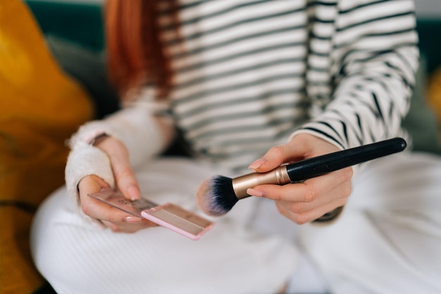 Colpo ritagliato del primo piano di una giovane donna ferita irriconoscibile con il braccio rotto avvolto in una benda di gesso bianco che applica cosmetici con pennello per il trucco a casa