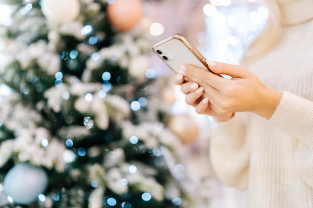 Colpo ritagliato del primo piano di una giovane donna irriconoscibile che utilizza lo smartphone digitante in piedi nella sala del centro commerciale celebra la vigilia di Natale