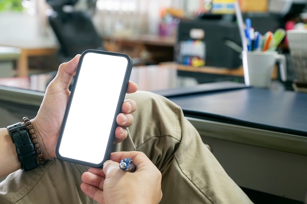 Colpo ritagliato della mano dell'uomo che tiene lo smartphone a schermo vuoto mentre è seduto al tavolo in ufficio.