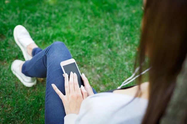 Colpo ritagliato di giovane ragazza bruna in cuffia che naviga in Internet sul suo smartphone nel parco. Rilassarsi su un prato verde. Concetto di stile di vita e tecnologia