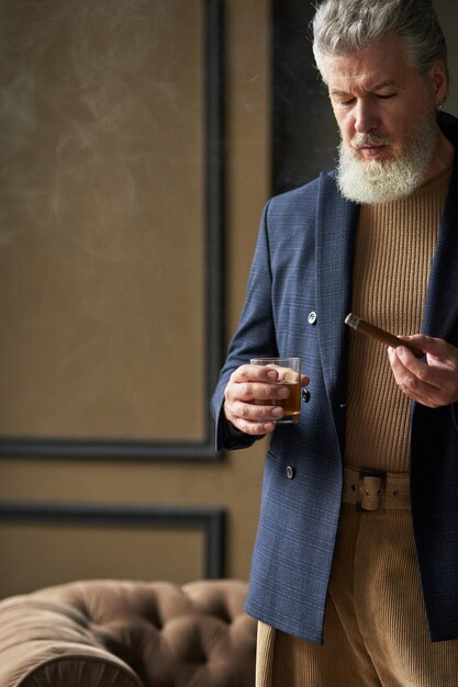 Colpo ritagliato di un uomo maturo serio con la barba che guarda il sigaro mentre lo tiene in mano e