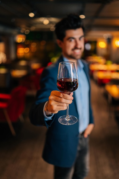 Colpo selettivo verticale di un uomo elegante positivo in abito di moda che tiene bicchieri di vino rosso in piedi nel ristorante sorridendo guardando la macchina fotografica