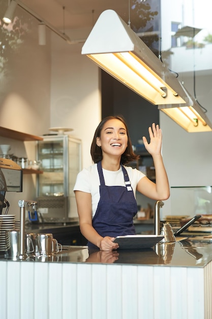 Colpo verticale di felice giovane barista asiatico lavoratore caffè onde al cliente riceve ordini dietro counte