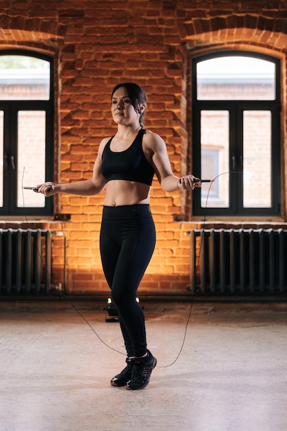 Colpo verticale di fitness giovane donna atletica con un bel corpo forte in abbigliamento sportivo che si esercita con la corda per saltare durante l'allenamento. Allenamento femminile caucasico muscolare fuori nella palestra scura.