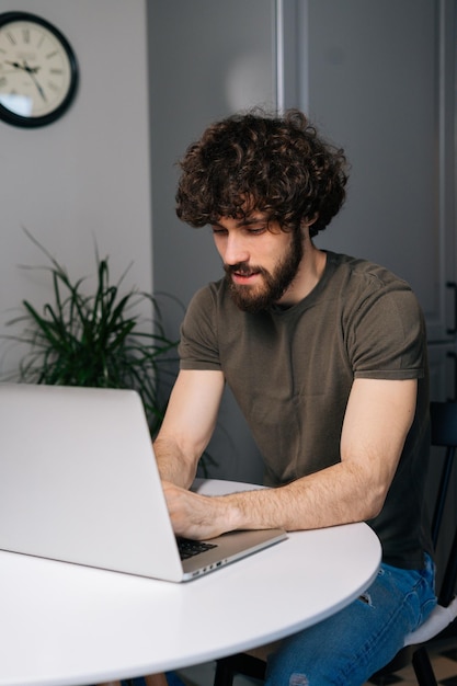 Colpo verticale di un bel giovane uomo d'affari barbuto che utilizza la tastiera del laptop per scrivere e-mail o un messaggio seduto al tavolo nella stanza della cucina