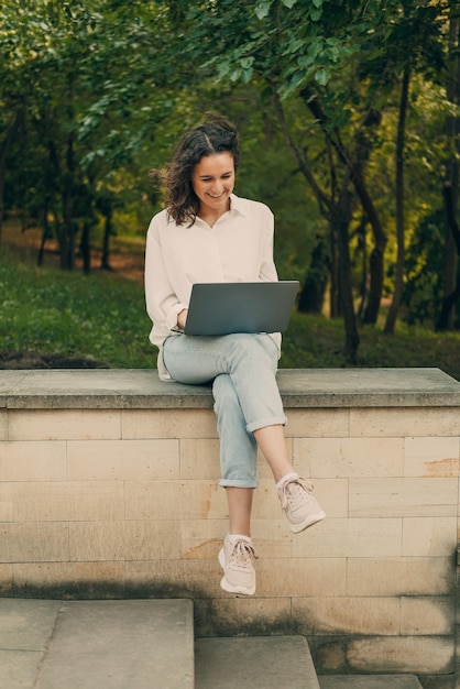 Colpo verticale di una donna che lavora al computer portatile mentre si siede sulla ringhiera di pietra