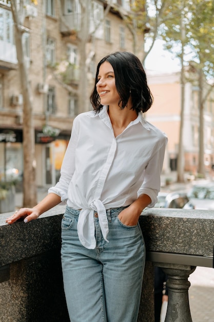 Colpo verticale di una donna felice che indossa una camicia bianca appoggiata alla ringhiera di pietra