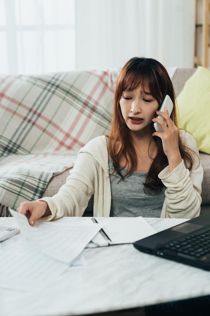 colpo verticale infelice donna asiatica sta guardando il conto mentre ha una lite al telefono mentre presenta la tassa al tavolo nel soggiorno di casa.