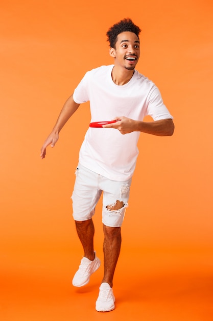 Colpo verticale integrale felice, carismatico ragazzo sportivo afroamericano come giochi attivi all'aperto, lancio del frisbee verso un amico e sorridente, godendo di una bella giornata di sole estivo, muro arancione