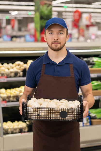 Colpo verticale medio lungo del moderno lavoratore del negozio che indossa una scatola di contenimento uniforme piena di funghi freschi