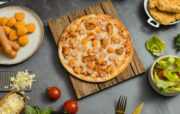 Colpo vista dall'alto di deliziosa gustosa pizza di formaggio con salsiccia a fette succosa posta su tavola di legno intorno con altri ingredienti di condimento di pollo fritto croccante fast food e verdure biologiche crude fresche