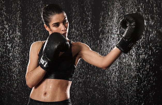 Combattente femminile 20s in abbigliamento sportivo facendo esercizi sportivi o praticando in guantoni da boxe neri mentre lancia pugni sotto le gocce di pioggia, isolato su sfondo scuro