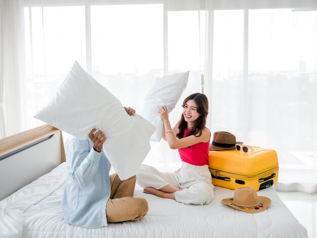 Combattimento di cuscini di coppia Bella giovane donna asiatica e uomo in camicia di jeans seduto e in lotta insieme ai cuscini vicino alla valigia gialla sul letto nella luminosa camera d'albergo Buone vacanze