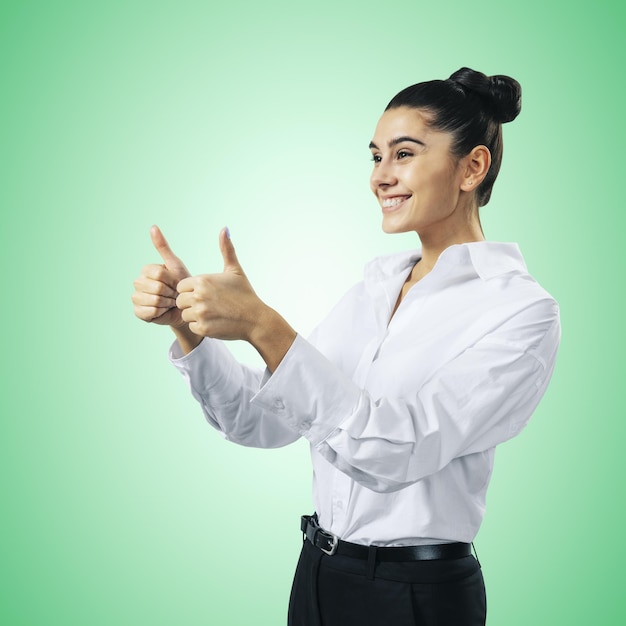 Come il concetto con felice giovane imprenditrice in camicia bianca che tiene i pollici in su su sfondo verde chiaro vicino isolato