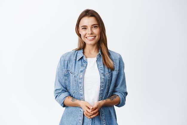 Come posso aiutarti Sorridente giovane donna con i capelli biondi tenendo le mani insieme e guardando amichevole alla fotocamera felice emozioni sfondo bianco