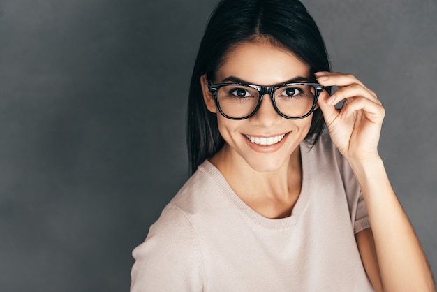 Come posso aiutarti? Vista dall'alto di una giovane donna attraente che si aggiusta gli occhiali e guarda la telecamera con un sorriso mentre è in piedi su uno sfondo grigio