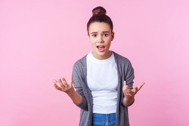 Come potresti Ritratto di ragazza adolescente bruna con acconciatura da panino in abiti casual alzando le mani chiedendo perché guardando indignato scontento facendo affermazioni indoor studio girato isolato su sfondo rosa