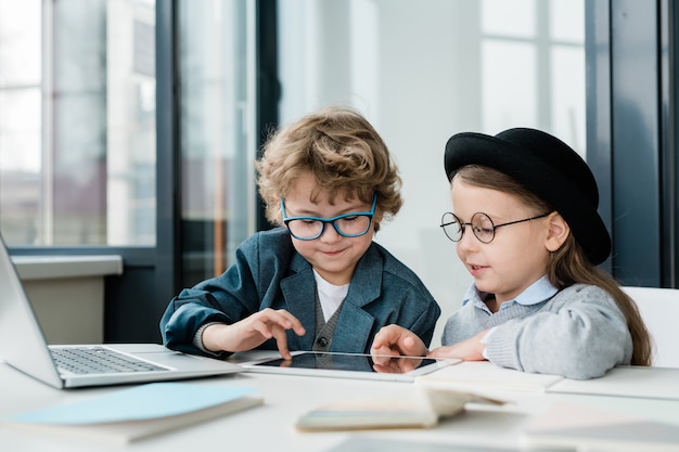 Compagni di scuola elementare carino in abbigliamento casual e occhiali da vista utilizzando la tavoletta digitale durante il lavoro di squadra a lezione