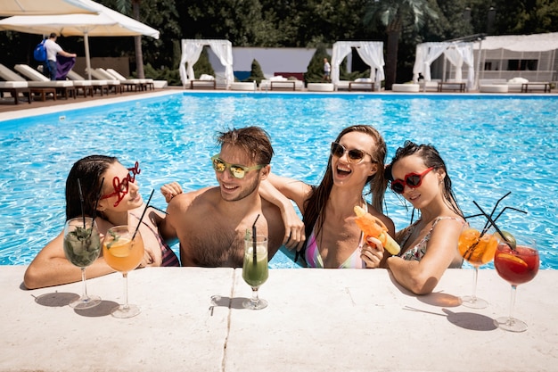 Compagnia di giovani ragazze e ragazzi allegri che si rilassano in piscina bevendo cocktail e giocando con pistole ad acqua all'aperto in una soleggiata giornata estiva vicino alla zona lounge