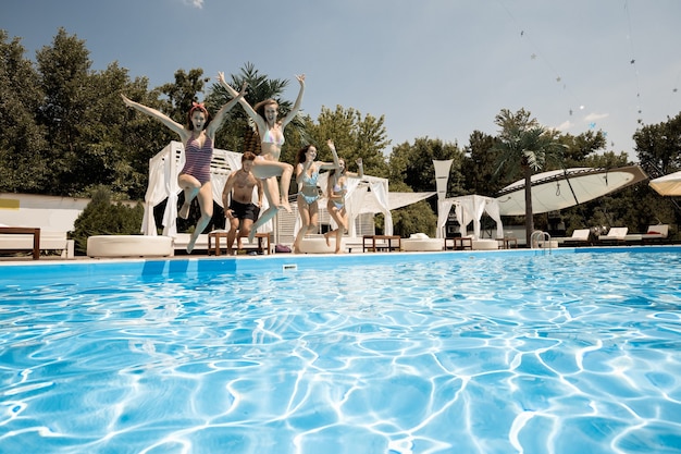 Compagnia di giovani ragazze e ragazzi allegri saltano insieme nella piscina accanto alla zona lounge all'aperto in una soleggiata giornata estiva