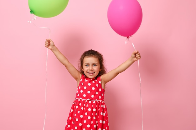 Compleanno Bambina vestita in abito rosa con motivo a pois alzando le braccia con palloncini colorati in mano, sorridente guardando la telecamera, isolata su sfondo rosa con spazio copia