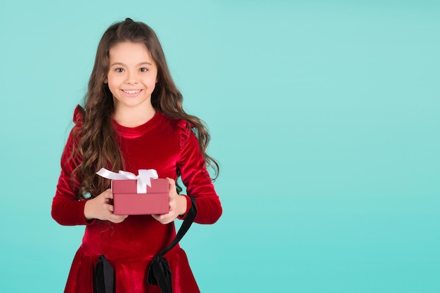 Compleanno, Capodanno, vacanze di Natale. Bambino felice in abito rosso tenere la casella su sfondo blu. Concetto di giorno di Santo Stefano. La ragazza con i capelli lunghi del brunette sorride con il presente. Fare regali, festeggiare, copiare lo spazio