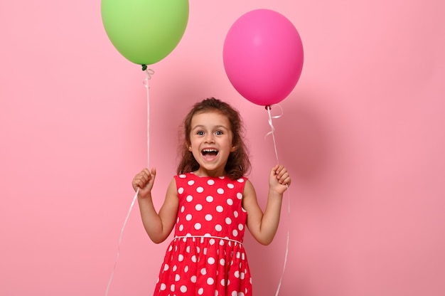 Compleanno ragazza vestita in abito rosa con palloncini colorati nelle sue mani, sorridente guardando la telecamera, esprimendo felicità, isolata su sfondo rosa con spazio copia. Concetti di infanzia felice