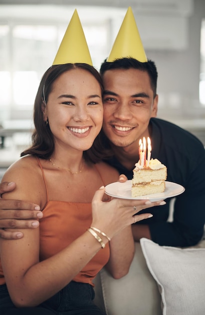 Compleanno ritratto e torta con una coppia nella loro casa che tiene il dessert per festeggiare in cappelli da festa Candela d'amore o romanticismo con un giovane uomo e una donna che festeggiano insieme nella loro casa