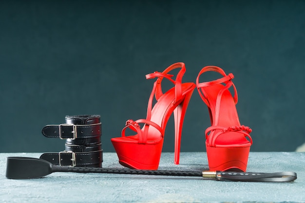 Completo BDSM per giochi di sesso per adulti. Scarpe rosse di striptease col tacco alto e manette, frusta su sfondo scuro - immagine