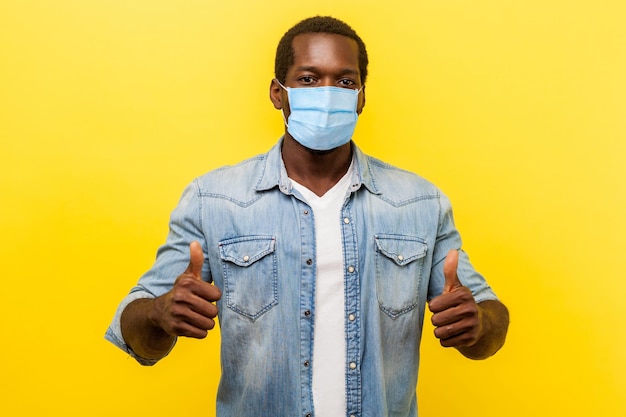 Complimenti, ottimo lavoro. Ritratto di un bell'uomo entusiasta in camicia casual in denim con maschera medica chirurgica che sorride e mostra come un gesto alla macchina fotografica. girato in studio isolato su sfondo giallo