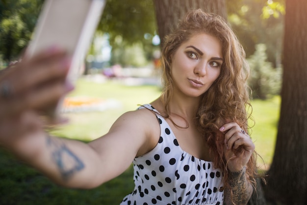 Comportamento da hipster. Stile giovanile moderno. Selfie di social media alla moda, sfondo del parco. Stile di vita estivo, ragazza bella ed elegante con il cellulare