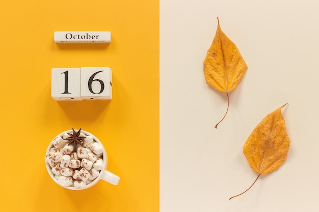 Composizione autunnale. Calendario in legno 16 ottobre, tazza di cacao con marshmallow e foglie autunnali gialle su fondo beige giallo. Vista dall'alto Flat lay Mockup Concept Ciao settembre.