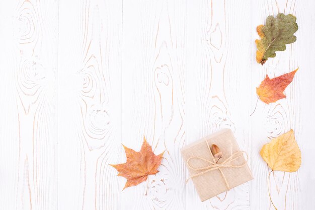Composizione autunnale con foglie secche autunnali un regalo avvolto in carta kraft steso in un angolo su uno sfondo di legno bianco rustico Spazio copia piatto