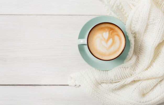 Composizione cappuccino invernale. Vista superiore della tazza di caffè blu con schiuma e maglione lavorato a maglia al tavolo in legno bianco. Concetto di bevande calde di riscaldamento