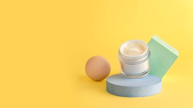 Composizione da scatola cosmetica aperta con crema che rimane sul podio geometrico. Sfondo giallo, grande striscione con spazio per la copia.