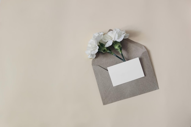 Composizione di cancelleria di compleanno di matrimonio femminile Mockup di invito biglietto da visita vuoto con busta artigianale isolata su sfondo tavolo beige Fiori di garofano bianco Vista dall'alto piatta