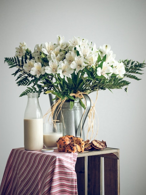 Composizione di cupcakes e bottiglia di latte posizionati vicino a bouquet di fiori bianchi su scatola di legno con panno su sfondo bianco