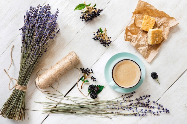 Composizione di fiori di caffè, biscotti, bacche e lavanda, vista dall'alto su legno bianco. Tazza blu con schiuma cremosa