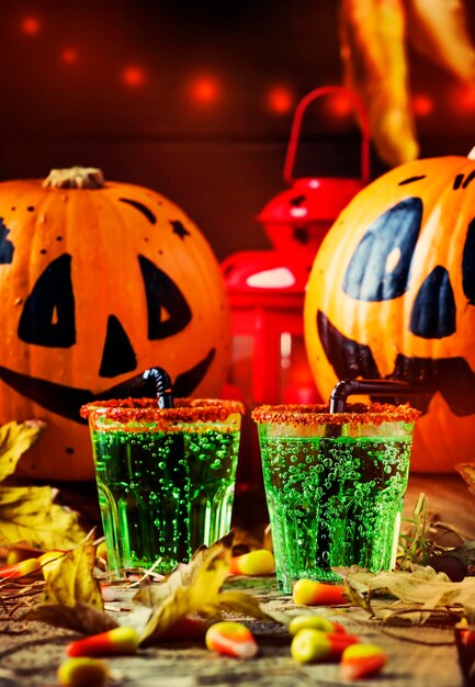 Composizione di Halloween con bevanda verde festiva e zucche beventi sorridenti con paglia di mais dolce e foglie cadute su sfondo scuro fuoco selettivo