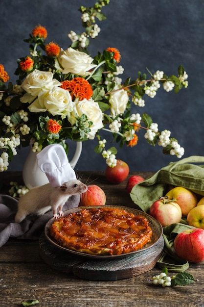 Composizione di natale. Il ratto è un cuoco vicino alla torta di mele e sul tavolo c'è un mazzo di fiori e mele