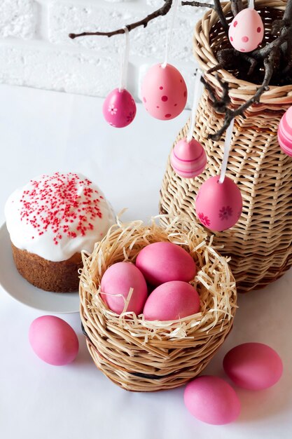 Composizione di Pasqua con i rami degli alberi decorati in un vaso di vimini, uova colorate di rosa nel cesto di vimini e torta di Pasqua su sfondo bianco