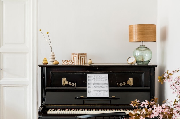 Composizione elegante all'interno del soggiorno con pianoforte nero, fiori secchi in vaso, orologio d'oro, lampada di design, scatole, spazio copia ed eleganti accessori personali in un arredamento moderno.