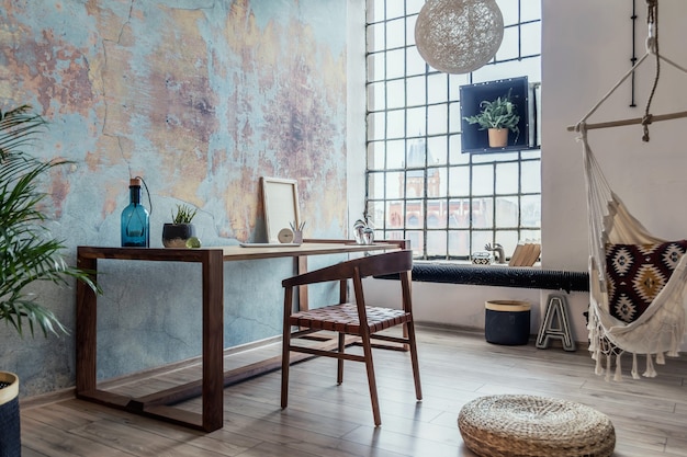 Composizione elegante e moderna degli interni dell'area di lavoro creativa con tavolo e sedia in legno, piante e accessori. Ampia camera con pareti creative e pavimento in parquet.
