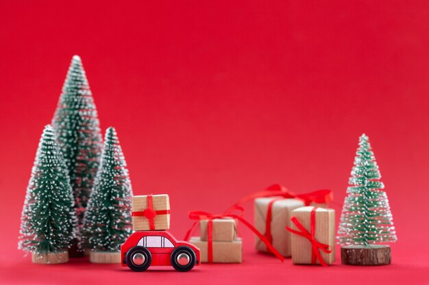 Composizione festiva Automobile rossa di Natale con una confezione regalo in una foresta di pini dove molti regali diversi su sfondo rosso. Felice anno nuovo concetto di carta.