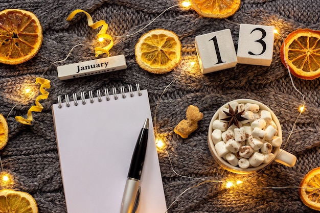 Composizione invernale. Calendario in legno 13 gennaio Tazza di cacao con marshmallow, blocco note aperto vuoto con penna