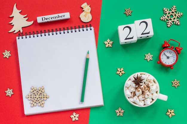 Composizione invernale. Calendario in legno 22 dicembre Tazza di cacao con marshmallow, blocco note aperto vuoto con matita, fiocco di neve, sveglia su sfondo rosso e verde. Vista dall'alto Mockup piatto