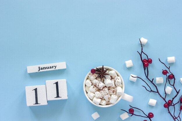 Composizione invernale. Cubi calendario in legno bianco. Dati 11 gennaio. Tazza di cacao, marshmallow e ramo decorativo con bacche rosse su sfondo blu Vista dall'alto Spazio di copia piatta