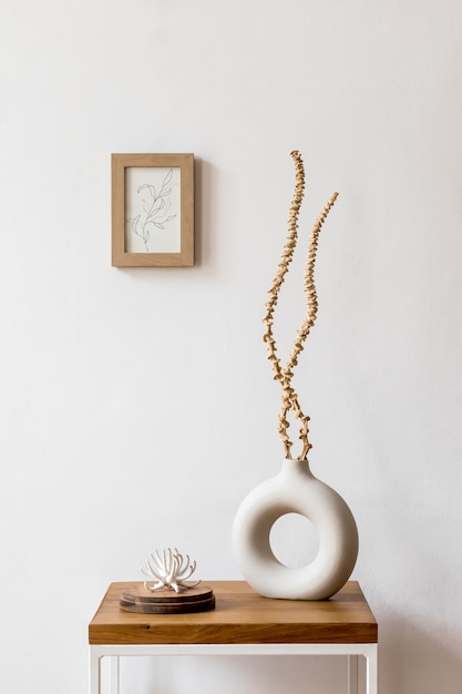 Composizione minimalista e di design di fiori secchi in vaso elegante, tavolino in legno, decorazione, cornice per foto e accessori all'interno bianco del soggiorno.