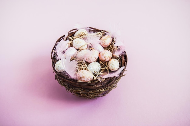 Composizione pasquale con decori tradizionali. Piccole uova colorate decorative e piume morbide in un cesto di vimini su sfondo rosa chiaro.