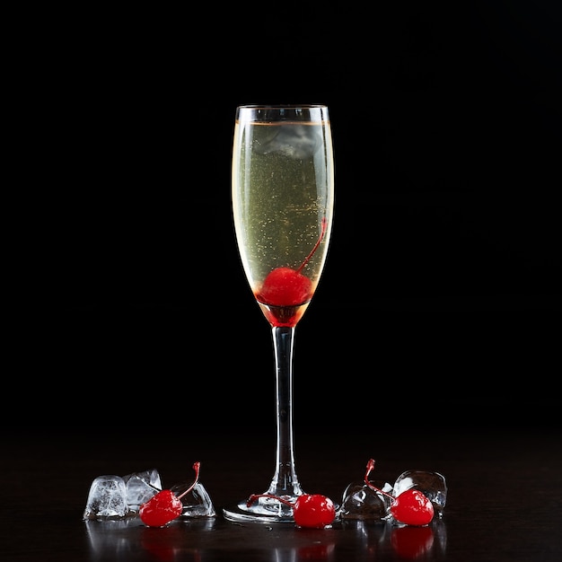 Composizione semplice di alto ed elegante bicchiere da cocktail in cristallo con bevanda fredda rinfrescante champagne trasparente, ciliegie rosse mature e cubetti di ghiaccio isolati su sfondo nero. Proporzioni immagine 1:1